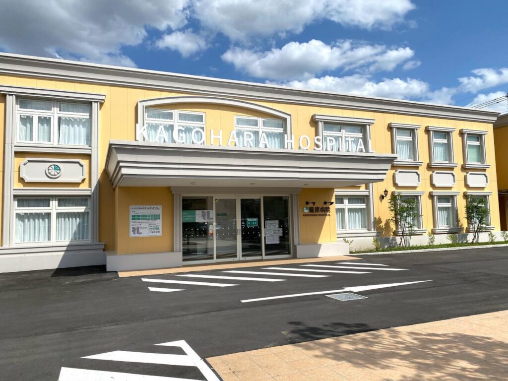黄色い壁の介護施設の玄関・車寄せ。晴天下、施設名が掲示されている。